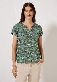 Haut vert à manches courtes avec un col en V, orné d'un motif rayé zèbre et de quatre boutons au niveau du col, associé à un pantalon beige.