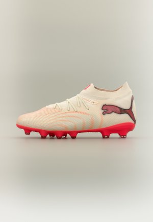 Crampon de football beige clair avec semelle rouge et logo Puma rouge sur le talon, motif à rayures orange, et fermeture à lacets avec col de cheville semblable à une chaussette.