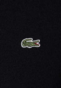 Svart stickad tyg med en liten, broderad grön krokodil-logotyp med röda detaljer, omlott i vitt. Diagonal ribbad struktur synlig.