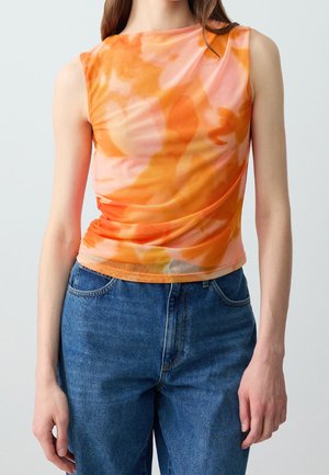 Kobieta ubrana w pomarańczowo-różowy top bez rękawów w stylu tie-dye oraz niebieskie dżinsy z wysokim stanem na jednolitym jasnoszarym tle.