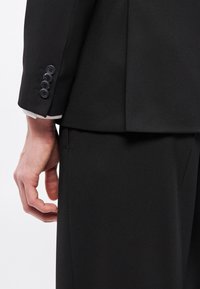 Czarna, dopasowana marynarka garniturowa z mankietem na cztery guziki, usztywnionym kołnierzem i gładką fakturą. Zbliżenie pokazuje część dłoni i detale spodni.