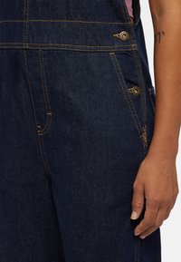 Right hand resting beside dark blue denim overalls with contrast orange stitching and metal buttons on hip and pocket area.