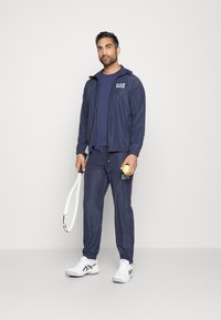 Navy sportswear with zip jacket and pants, featuring logo accents. Model holds a tennis racket and ball, wearing white shoes.
