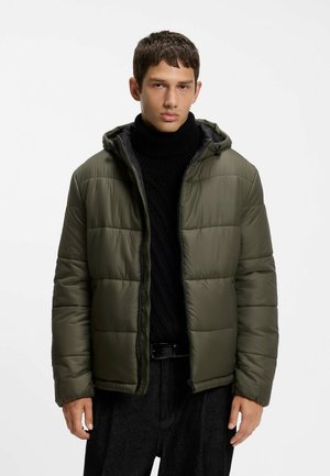 Junger Mann trägt eine olivgrüne Daunenjacke über einem schwarzen Rollkragenpullover mit schwarzen Hosen, steht vor einem schlichten weißen Hintergrund.
