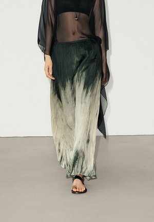 Femme portant un haut noir transparent et un pantalon large tie-dye noir et beige, marchant sur un sol gris avec des sandales noires.