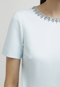 Light blue short-sleeve top featuring a round neckline embellished with silver gem-like accents, made from smooth fabric with a slight sheen.