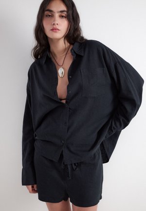 Kvinde med langt mørkt hår iført løs sort skjorte med knapper og matchende shorts, accessoriseret med en stor vedhæng halskæde.