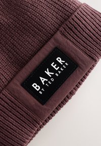 Bourgognefarvet strikket hue med ribbet tekstur; har en sort rektangulær etiket med teksten "BAKER BY TED BAKER" i hvidt.