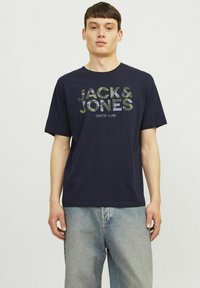 Tricou din bumbac bleumarin cu mâneci scurte, având un imprimeu mare cu logo-ul de camuflaj "JACK & JONES" și textul "SINCE 1990" pe față.