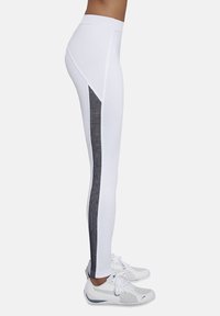 Białe legginsy z wysokim stanem mają czarną siateczkową wstawkę po zewnętrznej stronie. Połączone z białymi butami sportowymi.