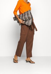Maglietta arancione a maniche lunghe abbinata a pantaloni cargo marroni. Il modello tiene in mano una borsa a tracolla in tessuto scozzese con motivi marroni, bianchi e neri.