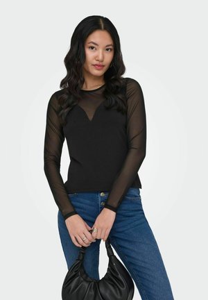 Femeie cu păr lung și negru purtând un top negru transparent cu mâneci lungi și blugi albaștri, ținând o geantă de piele neagră în fața ei.