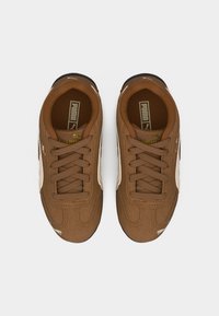 Ζευγάρι καφέ αθλητικά παπούτσια Puma με λευκές ρίγες στο πλάι και κορδόνια, σε θέα από πάνω πάνω σε απλό ανοιχτό φόντο.