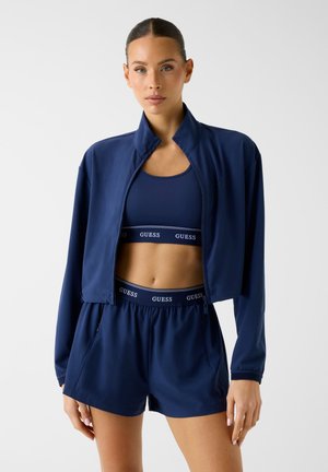 Donna che indossa una giacca sportiva blu navy, reggiseno sportivo e pantaloncini con il logo "GUESS" visibile sulle fasce in vita, in piedi davanti a uno sfondo semplice.