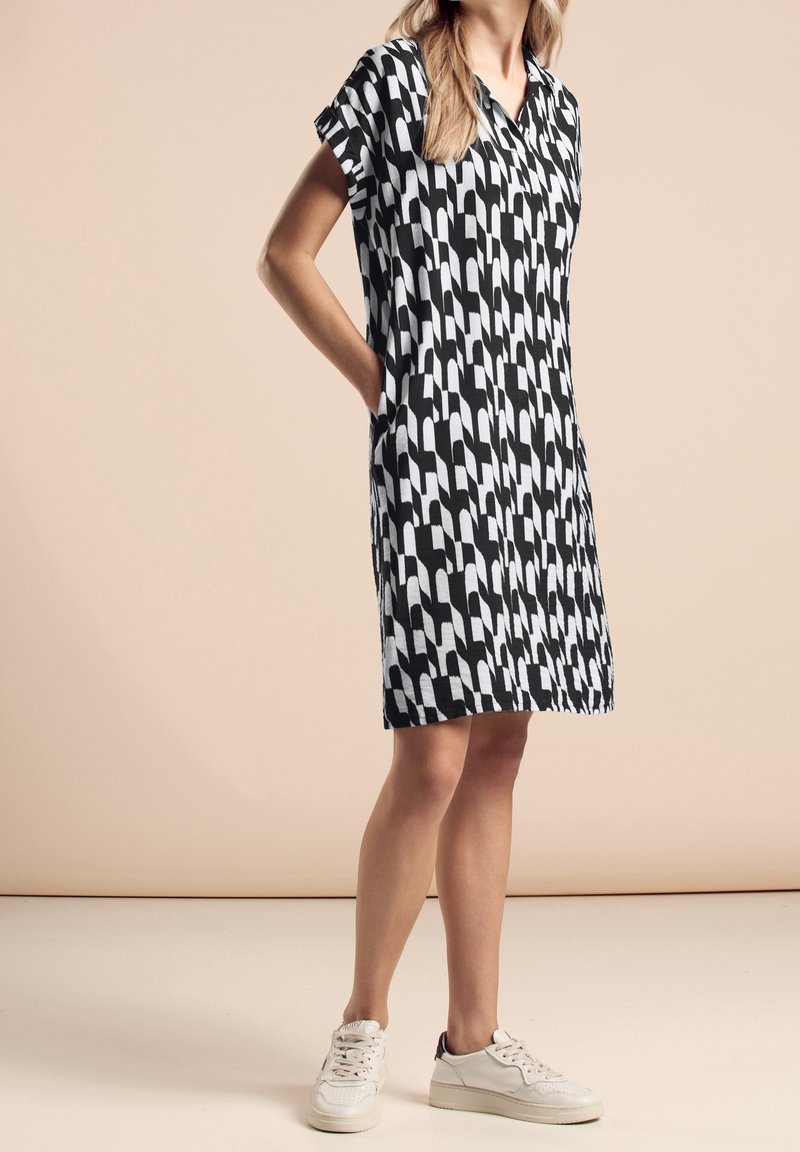 Mustavalkoinen kuvioitu mekko, jossa on lyhyet hihat, kaulus ja taskut, valmistettu kevyestä kankaasta, käytetään valkoisten lenkkarien kanssa.