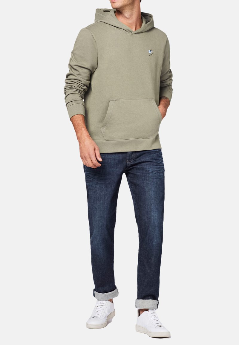 Olivengrøn hoodie med kængurulomme og ribbede manchetter, parret med mørkeblå jeans og hvide sneakers. Simpelt logo på brystet.