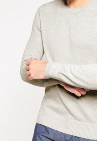 Pull en tricot gris clair avec un col rond, des poignets et un ourlet côtelés. Tissu texturé et design ajusté visibles. Pas d'imprimés ni de motifs visibles.