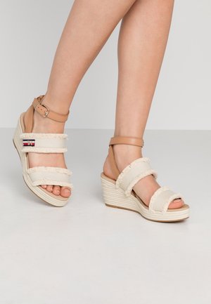 Sandały na koturnie z górą z kremowej tkaniny z postrzępionymi brzegami, opaskami na kostkę w odcieniu brązowym i plecioną lnianą podeszwą. Otwarty nos z widoczną stopą.