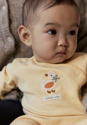 Bébé portant une tenue jaune avec un motif de canard et le texte « petit », regardant sur le côté en s’appuyant contre un tissu marron texturé.
