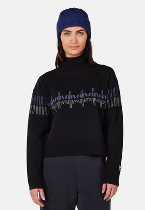 Schwarzer Strickpullover mit hohem Kragen, der horizontale Streifen aus blauen und silbernen Mustern aufweist. Kombiniert mit einer blauen Strickmütze.