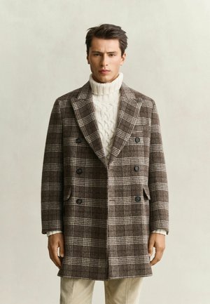 Mann trägt einen braun karierten doppelt geknöpften Mantel über einem weißen, kabelgestrickten Rollkragenpullover und steht vor einem einfarbigen Hintergrund.