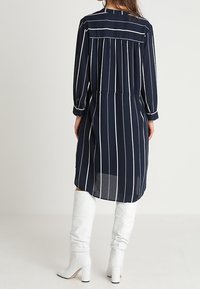 Marinblå klänning med vita vertikala ränder, lång ärm, elastisk midja och rygdrapering, kombinerad med knähöga vita klackstövlar.