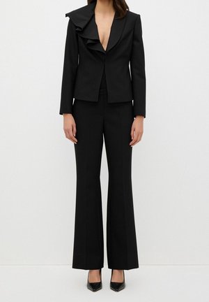 Femme portant un costume noir sur mesure avec un blazer à revers volanté et un pantalon à jambes larges, associé à des talons noirs à bout pointu.