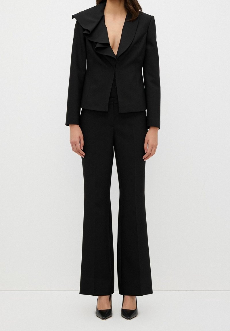 Femme portant un costume noir sur mesure avec un blazer à revers volanté et un pantalon à jambes larges, associé à des talons noirs à bout pointu.