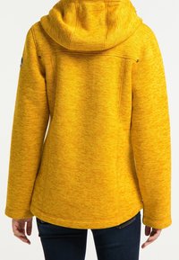 Žlutá kapucňová bunda vyrobená z texturované látky, která má volný střih, čisté švy a zaoblený lem se bočními kapsami.