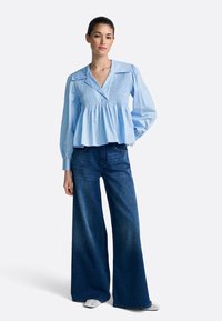 Blusa azul claro abotonada con mangas abullonadas y detalle fruncido, combinada con vaqueros de pierna ancha. Tela suave, silueta relajada.