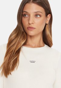 Top bianco a maniche lunghe con motivo a costine e scollo tondo, con un piccolo logo nero "GUESS JEANS" sul petto. Texture liscia.