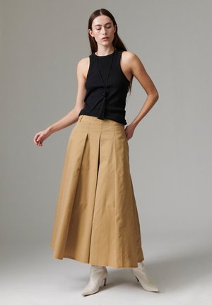 Femme en haut noir sans manches, longue jupe beige à larges jambes, bottes blanches à talons et collier noir à pampilles, debout devant un fond gris uni.