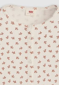 Weißes Baumwolloberteil mit rundem Ausschnitt, vertikaler Rippstruktur, kleinem Blumenmuster in Rostfarbe und fünf dekorativen Knöpfen in der Mitte.
