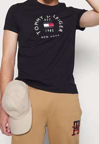 Camiseta de algodón negra con un logo redondo que presenta texto blanco y una pequeña bandera de color. Combinada con pantalones de chándal beige que muestran iniciales bordadas.
