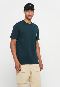 Jongeman in een donkergroene polo met zak, beige cargobroek en een zwarte honkbalpet, staande voor een effen witte achtergrond.