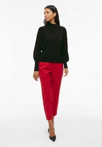 Pull noir avec manches bouffantes texturées, associé à un pantalon rouge ajusté et des escarpins noirs pointus. Design simple et moderne, sans motifs.