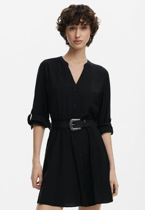 Schwarzes Knopf-up Kleid mit V-Ausschnitt und hochgerollten Ärmeln, tailliert mit einem strukturierten schwarzen Gürtel, der mit einem silbernen Schnallen-Detail versehen ist.