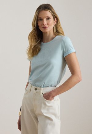 Hosszú szőke hajú nő, világoskék rövid ujjú inget visel, amelyet magasderekú szürke-fehér nadrágba tűrt be, kezét a zsebében tartja.