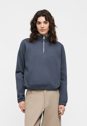 Vrouw met krullend haar die een marineblauwe sweatshirt met een kwart rits en kangoeroezak draagt en beige broek, staand tegen een effen achtergrond.