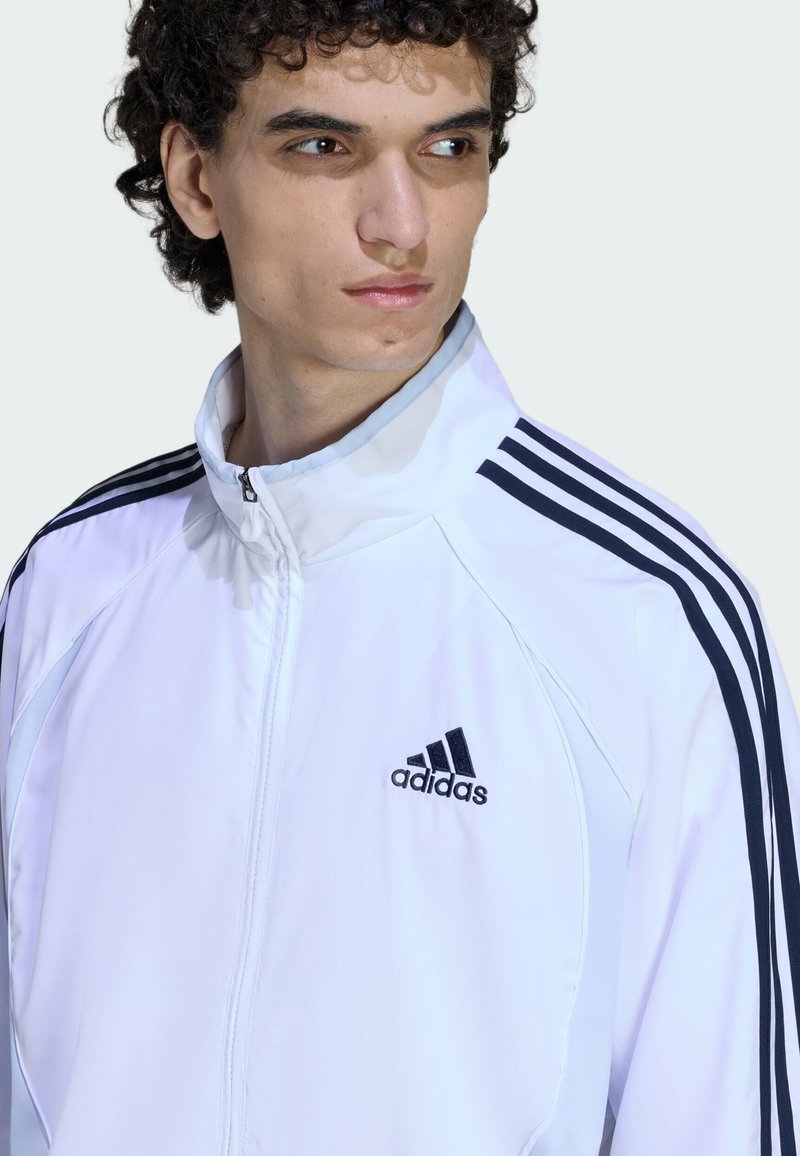 Veste de sport blanche avec fermeture éclair, motif trois bandes noires sur les épaules, et logo Adidas noir sur le côté gauche de la poitrine. Texture lisse.