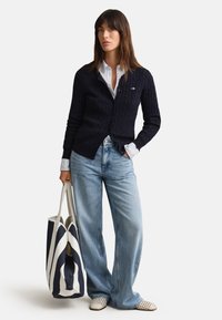 Donna che indossa un cardigan blu navy lavorato a trecce sopra una camicia bianca con jeans azzurri a gamba larga, tenendo una grande borsa a righe blu navy e bianca.