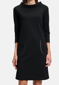 Svart klänning med en cowl-krage, trekvartsärmar, två stora påsydda fickor med kontrasterande kant och ett mjukt, texturerat tyg.