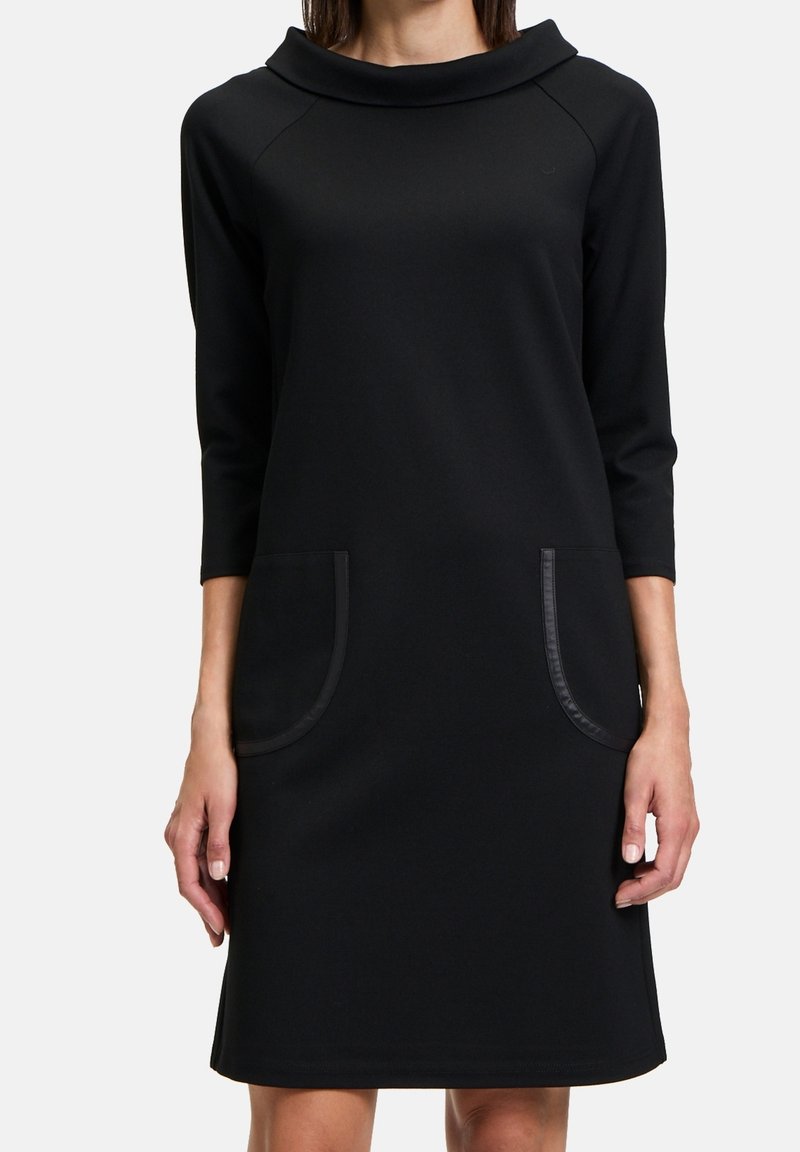 Svart klänning med en cowl-krage, trekvartsärmar, två stora påsydda fickor med kontrasterande kant och ett mjukt, texturerat tyg.