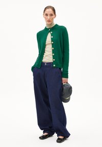 Vrouw in een groene cardigan met knopen over een beigekleurige coltrui, losse donkerblauwe broek, zwarte schoenen, die een donker baseballpet vasthoudt.