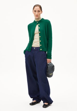 Vrouw in een groene cardigan met knopen over een beigekleurige coltrui, losse donkerblauwe broek, zwarte schoenen, die een donker baseballpet vasthoudt.