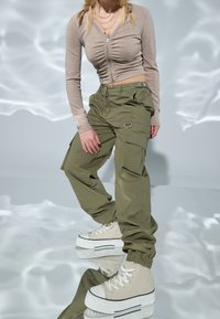Světle hnědý, dlouhý rukáv, zipový cropped top s nařasenými detaily, spárovaný s olivově zelenými cargo kalhotami s kapsami a manžetami na kotnících. Béžové platformové tenisky.