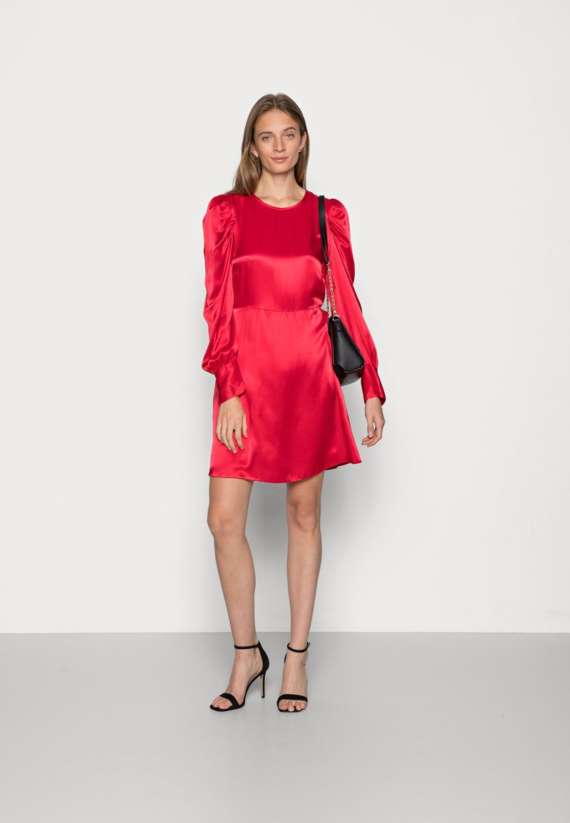 Rotes Satin-Kleid mit langen, puffigen Ärmeln, einem Rundhalsausschnitt und einem ausgestellt geschnittenen Saum. Kombiniert mit schwarzen Riemchenschuhen und einer kleinen schwarzen Handtasche.