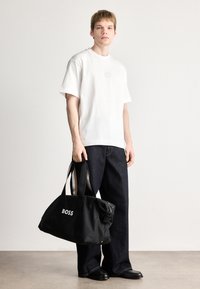 Λευκό κοντομάνικο μπλουζάκι με ανάγλυφο λογότυπο, συνδυασμένο με σκούρο μπλε φαρδύ τζιν παντελόνι και μια μαύρη νάιλον τσάντα duffel με λευκό λογότυπο.