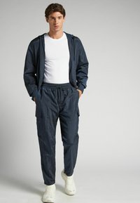 Marinblå nylon träningsoverall med zip-jacka och byxor med elastisk midja. Den har en huva, sidofickor och vita sneakers. Slät textur.
