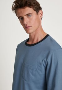 Blauwe lange mouwen shirt van zacht katoen. Heeft een zwarte halslijn en een klein zakje aan de linkerkant. Soepele textuur.